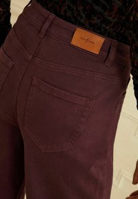 Burgundi värvi denimist teksased kitsas lõikes, millel on nahklabel vöö kohal ja tagatasku, millel on diskreetne logo tikand.