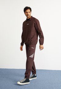 Vīrietis iet iekštelpās, ģērbies tumši brūnā Adidas treniņtērpā ar baltām svītrām un melnajās augstajās Converse kedās uz zilā paklāja.