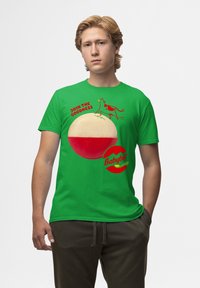 T-shirt in cotone verde con una grafica di una mucca e formaggio Babybel. Il testo recita "UNISCI ALLA BONTÀ" in rosso. Maniche corte e vestibilità rilassata.
