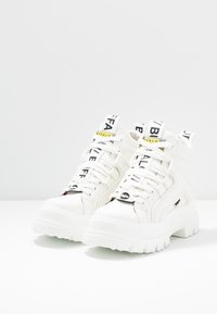 Sneaker alte bianche realizzate in materiale sintetico con suole spesse e testurizzate, dotate di cinturini e lacci brandizzati neri e gialli.