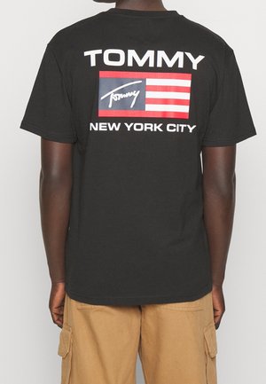 Osoba ubrana w czarną koszulkę z białym napisem "TOMMY", czerwoną i białą flagą w paski oraz napisem "NEW YORK CITY" na plecach, w połączeniu z beżowymi szortami typu cargo.