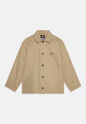 Giacca impermeabile beige con colletto, bottoni frontali, due tasche laterali e polsini elastici. Presenta una piccola patch con logo sul petto.