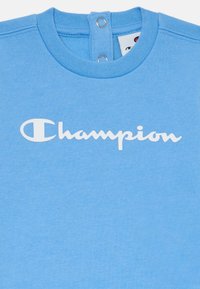 Svetlo modra majica s krojem, z belim logotipom "Champion". Dva pritisna gumbka na hrbtni strani vratu. Mehak tekstil.