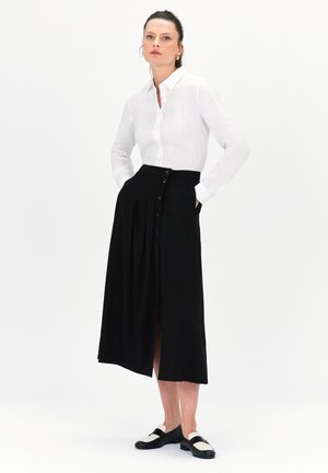 Camicia bianca in lino con bottoni, gonna midi nera plissettata con spacchi laterali e dettagli in bottone dorato, abbinata a mocassini bianco e neri.