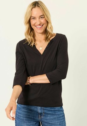 Usmievavá žena s blond vlasmi má na sebe čiernu blúzku s dlhým rukávom a modré džínsy, pózuje s jednou rukou založenou a druhou vystretou vpred.