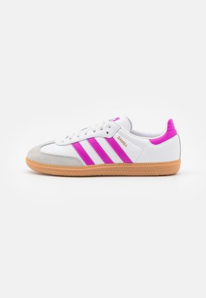 Basket blanche Adidas Samba avec des rayures roses, languette rose au talon, bout en daim beige et semelle en gomme sur fond blanc.
