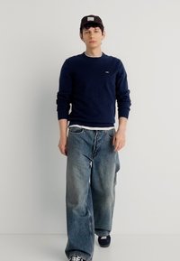 Νεαρός άνδρας που φοράει μαύρο καπέλο, μπλε σκούρο πουλόβερ, λευκό εσώρουχο, φαρδιά ξεθωριασμένα τζιν και μαύρα αθλητικά παπούτσια περπατώντας μπροστά σε απλό λευκό φόντο.