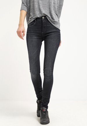 Personne portant un jean skinny foncé, un haut à manches longues gris et des baskets noires Reebok, debout devant un fond clair uni.