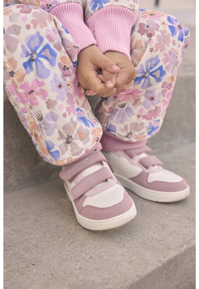 Rosa Hose mit Blumenmuster, elastische Bündchen und rosa Pulloverärmel. Hellrosa und weiße Sneaker mit Klettverschlüssen und strukturierten Akzenten.