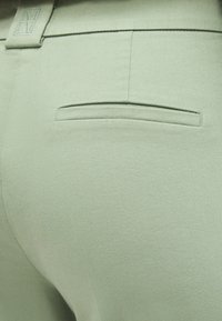 Εκλεπτυσμένη φωτογραφία μεσαιωνικής τσέπης πίσω σε ανοιχτό πράσινο εφαρμοστό παντελόνι, με θηλιά ζώνης που φέρει κεντημένο λογότυπο "MM".