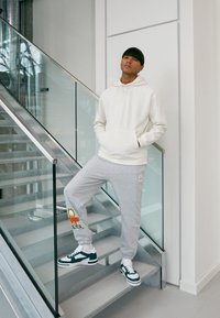 Sweat-shirt à capuche blanc, pantalon de survêtement gris avec un dessin animé, et baskets vertes et blanches, debout sur des escaliers modernes avec une rambarde en verre.