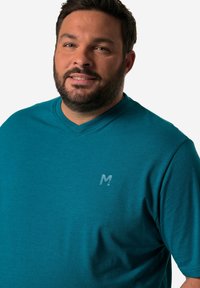 Tyrkysové tričko s výstřihem do V vyrobené z měkké látky, které má na hrudi malé, nenápadné logo. Design má krátké rukávy a volný střih.