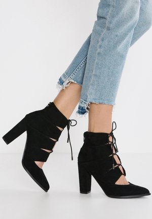 Femme portant un jean bleu clair à ourlet effiloché et des bottines noires en daim à lacets et talons hauts, debout sur un sol blanc.