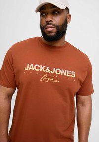 Pruun puuvillane t-särk, millel on suur heleda kreemika fondiga "JACK&JONES" tekst ja allkiri graafika. Lühikesed varrukad, ümar kaelus, sile tekstuur.
