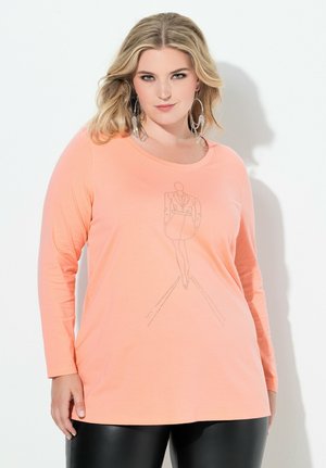 Woman with blonde hair wearing large silver hoop earrings, peach long-sleeve top with a minimalist blazer sketch, and black pants.