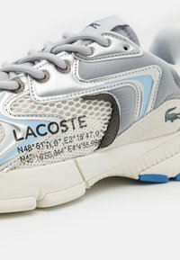 Tenisky Lacoste s horní částí z šedé a stříbrné síťoviny, modrými akcenty, texturovanou vrstvou a výrazným logem na boku. Bílá gumová podrážka.