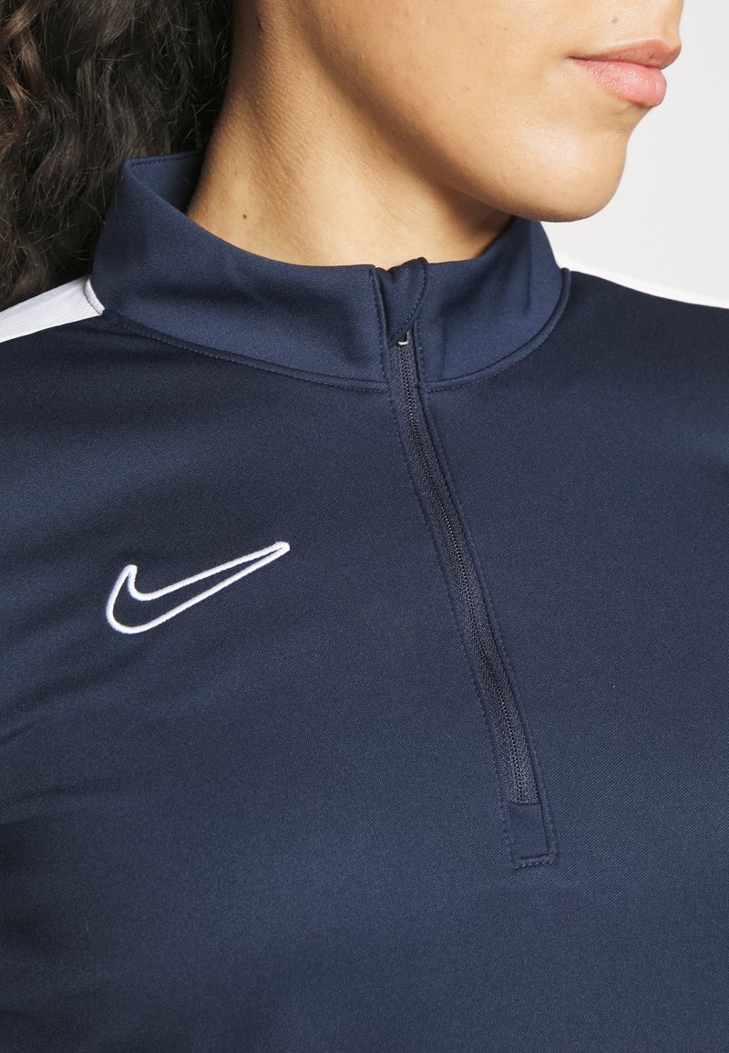 Nahaufnahme einer Person, die eine marineblaue Sportjacke mit Reißverschluss und einem weißen Nike-Logo auf der Brust trägt.