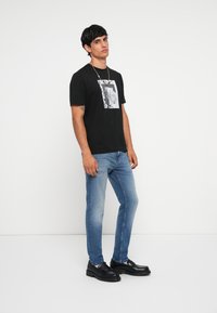 Black cotton t-shirt with a graphic print, paired with light blue denim jeans. Black leather shoes with a thick sole, standing against a white background.