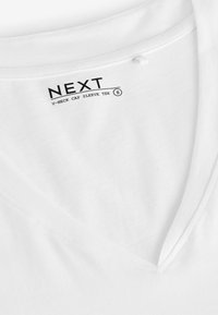 Bijela pamučna majica s V izrezom i rukavima na kapu. Ima glatku teksturu i otisnutu etiketu na kojoj piše "NEXT V-NECK CAP SLEEVE TEE S."