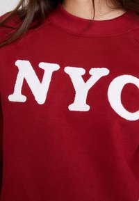 Maroon sweatshirt featuring large, white, plush letters spelling "NYC." Smooth fabric with ribbed collar. Close-up view of neckline and lettering.