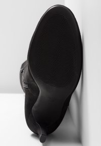 Botte noire en daim à talon haut avec semelle texturée et fermeture éclair latérale, inclinée contre une surface et un mur blancs.