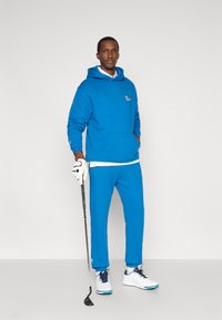 Zils džemperis ar kangaroo kabatu, atbilstošas zilas sporta bikses, baltā T-kreklā, baltas kurpes ar zilām akcentiem un golfa nūja rokā.