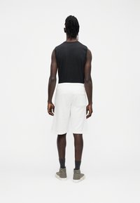 Schwarzes ärmelloses Oberteil, weiße Shorts, strukturierter Stoff, elastischer Bund, zwei Taschen. Sichtbare Knöchelsocken und legere Schuhe. Neutraler Hintergrund.