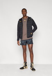 Μαύρο τζάκετ με φερμουάρ και μοτίβο σχήματος V, καφέ μπλούζα, μπλε σορτς με σχέδια και μαύρα μποτάκια με μπεζ λεπτομέρειες.