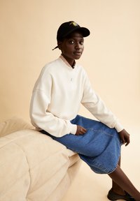 Witte sweatshirt met verlaagde schouders, gecombineerd met een blauwe denimrok. Zwarte pet met kleurrijke zon borduursels. Glad, neutraal achtergrond.