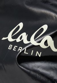 Tessuto nero testurizzato con il testo bianco in grassetto che recita "lala BERLIN". La superficie riflette la luce, creando una leggera lucentezza.