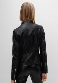 Kobieta o prostych blond włosach, ubrana w dopasowaną czarną skórzaną kurtkę i pasujące czarne skórzane spodnie, widziana z tyłu na tle jednolitym.