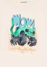 Krém színű ing, zöld színű szörnyautó grafikával, csillámos díszítéssel, nagy fekete kerekekkel és a "WOW!" kék stilizált szövegezéssel.