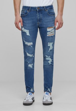 Jean skinny en denim bleu avec plusieurs patches déchirés, des accents de couleur plus claire et une silhouette ajustée. Taille réglable.