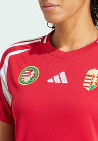 Camiseta deportiva roja con franjas de acento blancas, logo bordado de la federación húngara de fútbol y logo de Adidas en blanco. Textura de tela suave.