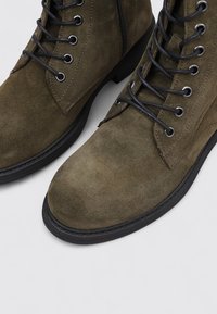 Botas de tobillo de ante verde oliva con cordones negros y ojales de metal, con punta redonda y suela de goma negra baja, que presentan una cremallera en el lateral.