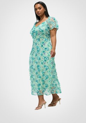 Kobieta w turkusowej, kwiatowej sukience midi z bufkami i złotych sandałach na wysokim obcasie, stojąca na jednolitym białym tle.