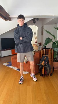 Noor mees musta jope ja beežide lühikeste pükstega seisab käed ristis elutoas, kus on oranž diivan, toataim ja puidust treeningseade.