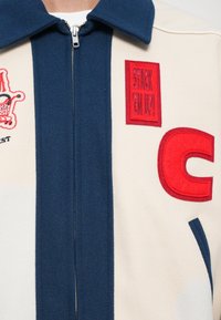 Creme- og marineblå fleecejakke med lynlåsfront. Har røde broderede lapper, herunder "STACK EM UP!" og et stort "C"-accent.