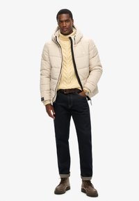Beige pufferjakke med en sort lynlås, over en crèmekabelstriktrøje, parret med mørke jeans og brune snørebåndsstøvler.