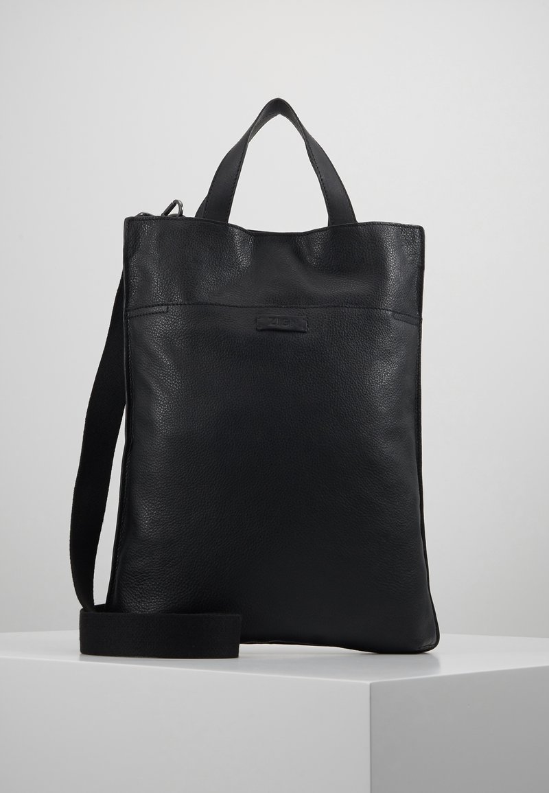 Sac cabas en cuir noir avec double poignées et bandoulière amovible, présenté sur une plateforme blanche sur fond neutre.