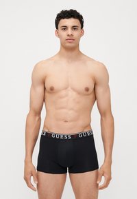 Boxeri negri din material elastic, cu un brâu gros cu logo-ul roșu și alb "GUESS". Țesătură netedă, design ajustat și croială de lungime medie.