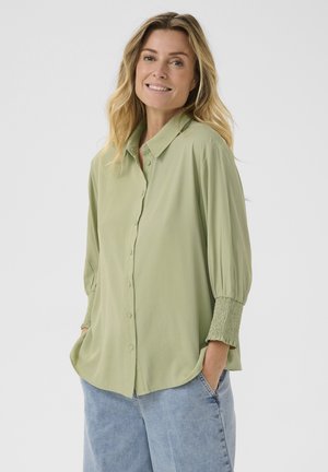 Blouse vert clair avec un col, fermeture à boutons et manches bouffantes avec des poignets smockés. Portée avec un jean bleu clair.