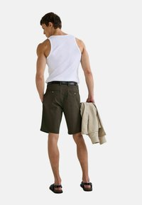 Modèle portant un débardeur blanc, un short vert olive et des sandales noires, tenant une veste beige clair. Comprend une ceinture noire et des poches arrière.