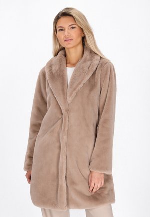 Donna che indossa un lungo cappotto in pelliccia sintetica beige con un ampio colletto, in piedi davanti a uno sfondo bianco semplice.