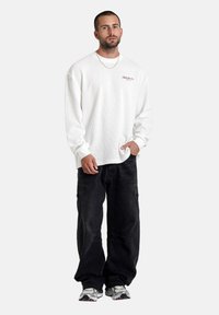 Man in een wit shirt met lange mouwen, zwarte baggy broek en grijze sneakers, staand met één hand in de zak tegen een witte achtergrond.