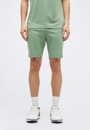 Mees kannab helerohelisi lühikesi pükse, sobivaid rohelist särk, valged keskmise pikkusega sokid ja valged spordijalatsid, seisab ühevärvilise tausta ees.