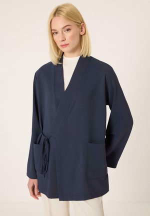 Ξανθιά γυναίκα που φοράει ένα ναυτικό μπουφάν τύπου wrap με πλαϊνές τσέπες και φαρδιά μανίκια πάνω από λευκό τοπ, σε απλό φόντο.