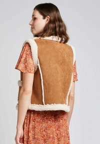 Gilet en suede marron avec des bordures en fausse fourrure crème, présentant un design ouvert et des accents d'épaule, associé à une robe orange à motifs.
