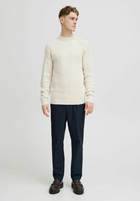 Maglione a coste color beige chiaro con colletto alto, abbinato a pantaloni blu scuro e mocassini neri chunky. Design minimalista, forma aderente.