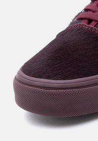 Κοντινή λήψη της μύτης ενός μπορντό αθλητικού παπουτσιού με υφή από καουτσούκ στη σόλα και επάνω μέρος που μοιάζει με γούνα, σε λευκό φόντο.